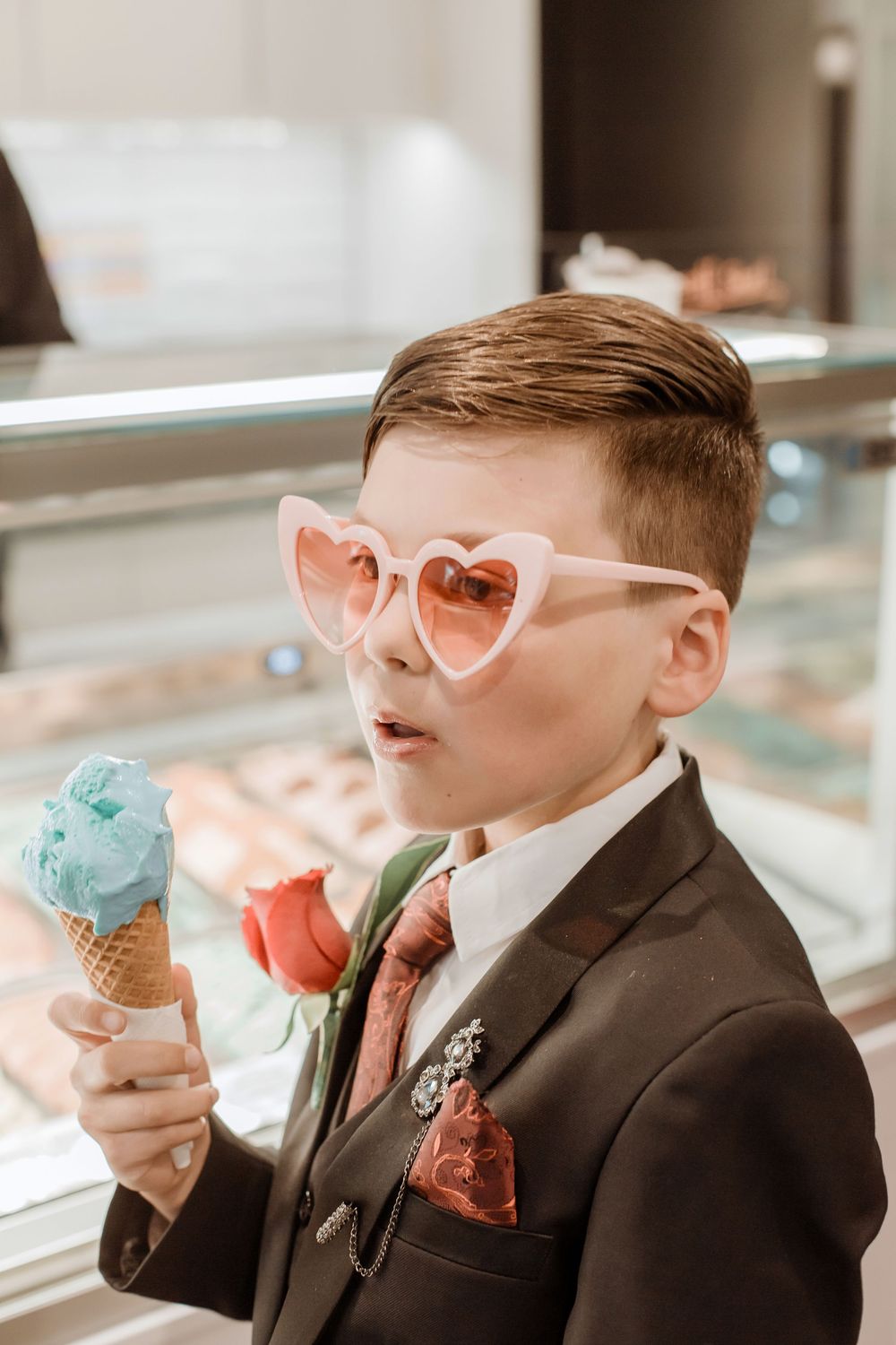 Young boy in a black suit enjoys blue ice cream cone while wearing heart-shaped pink sunglasses at a dessert counter.