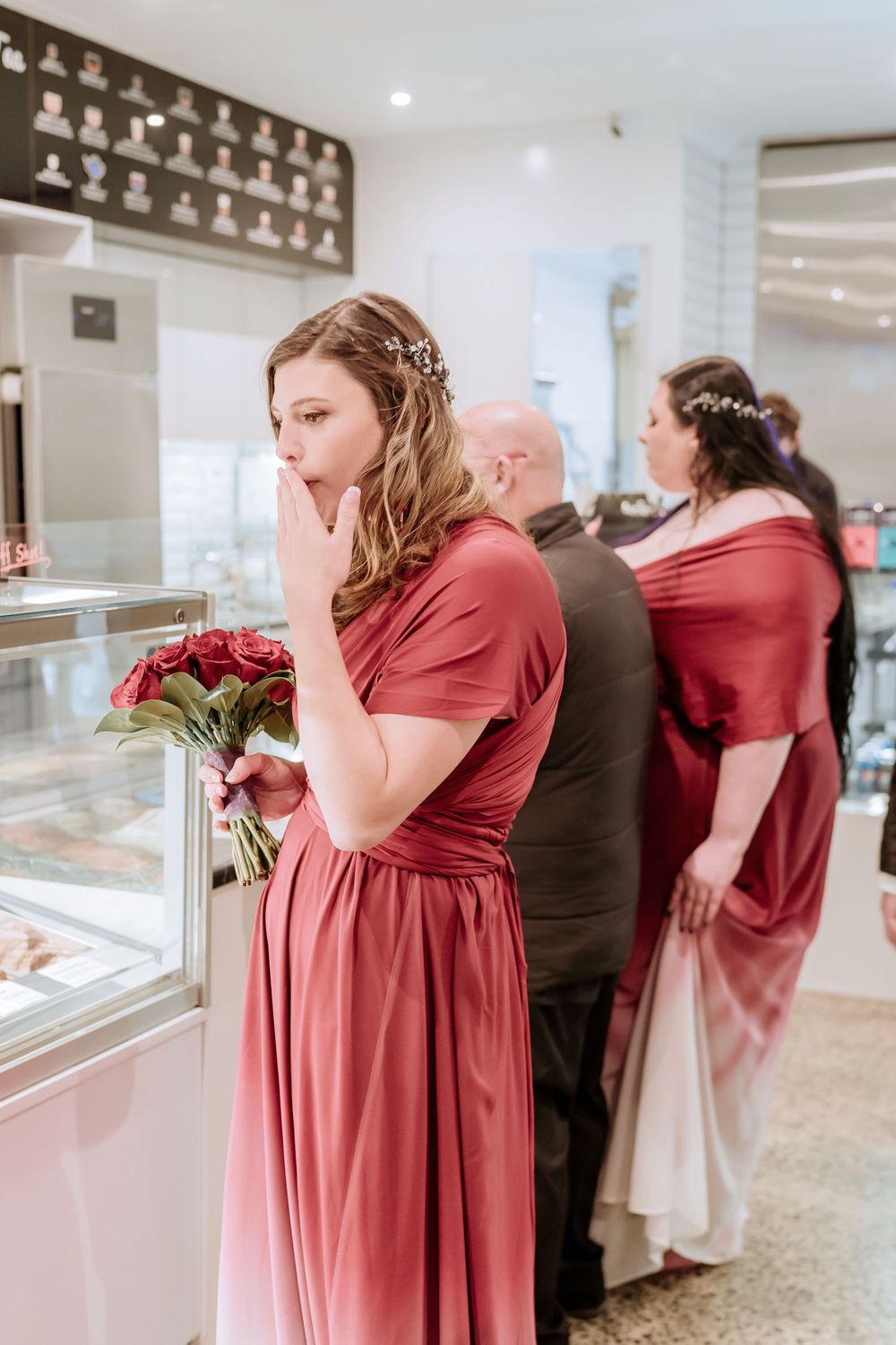 People wearing red bridesmaid dresses look into jewelry display cases at a shopping venue.