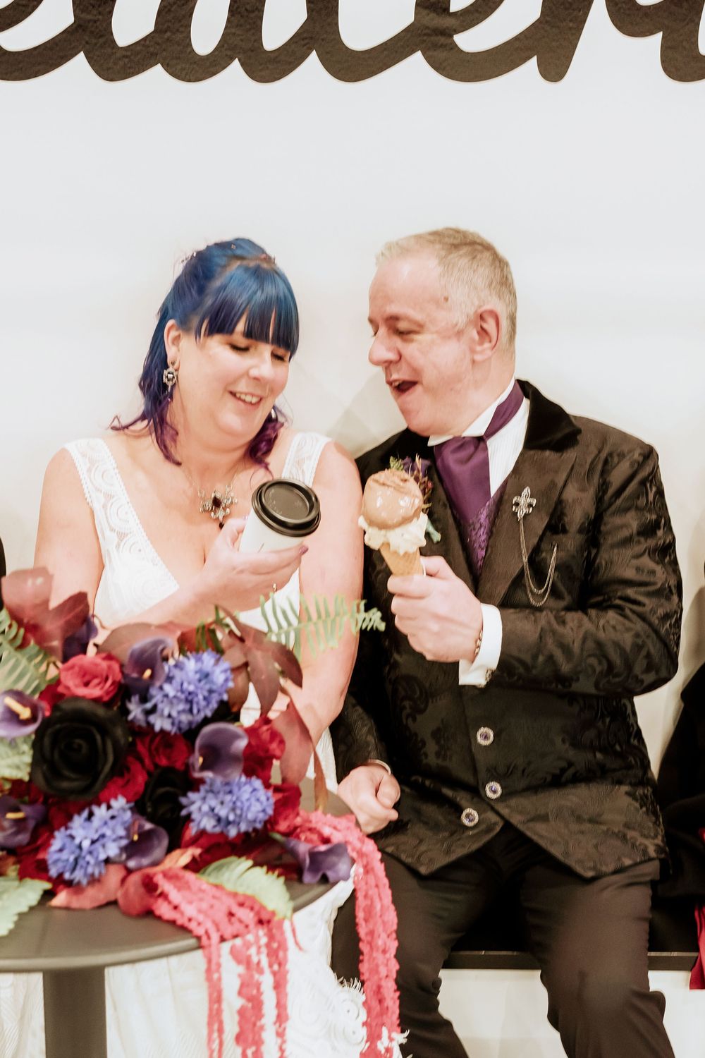 Newlywed couple sharing ice cream together at wedding reception with purple and red floral bouquet on table.