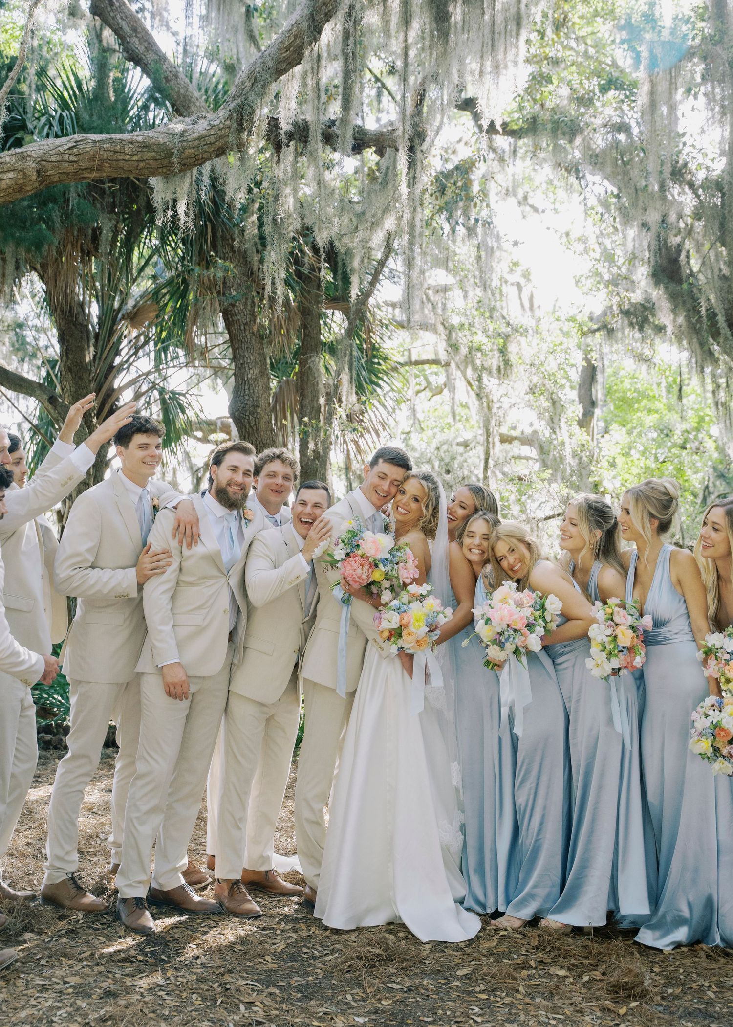 Wedding party poses under moss-covered oak trees, bridesmaids in light blue dresses holding bouquets.