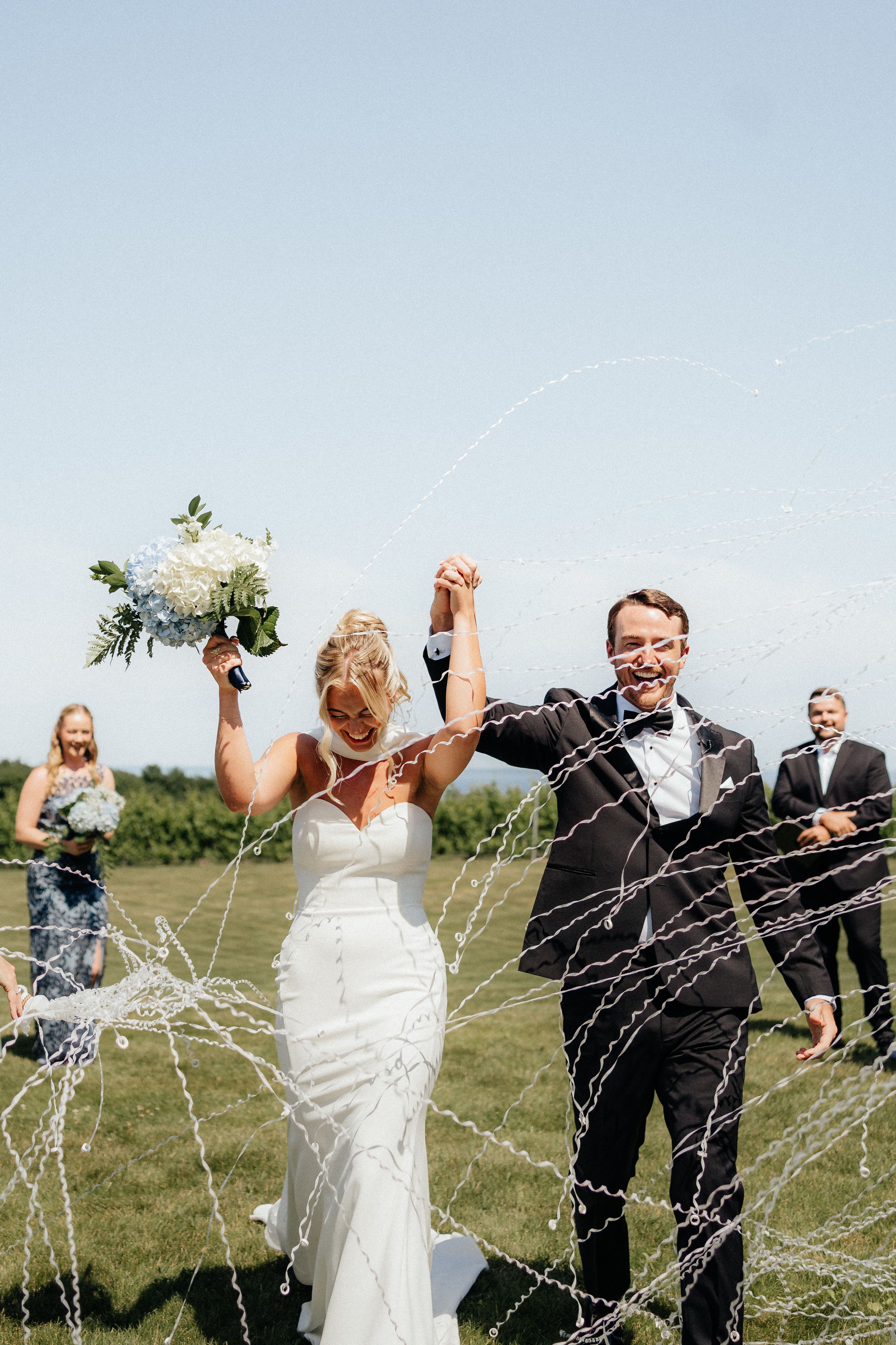 Wedding party celebrates by spraying champagne outdoors in a field during a joyful summer celebration.