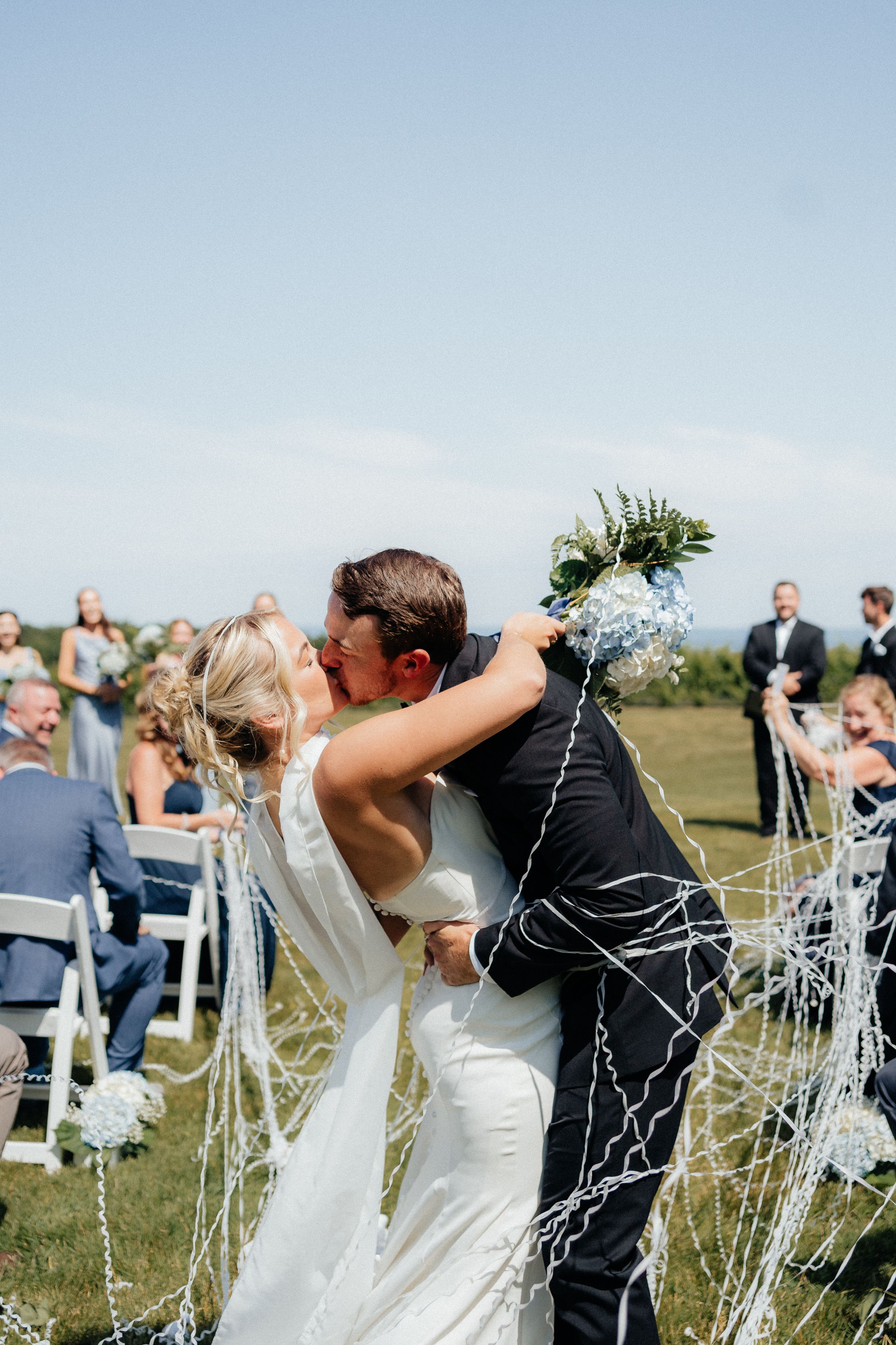 Wedding guests shower a couple with confetti during an outdoor ceremony celebration against a blue sky.