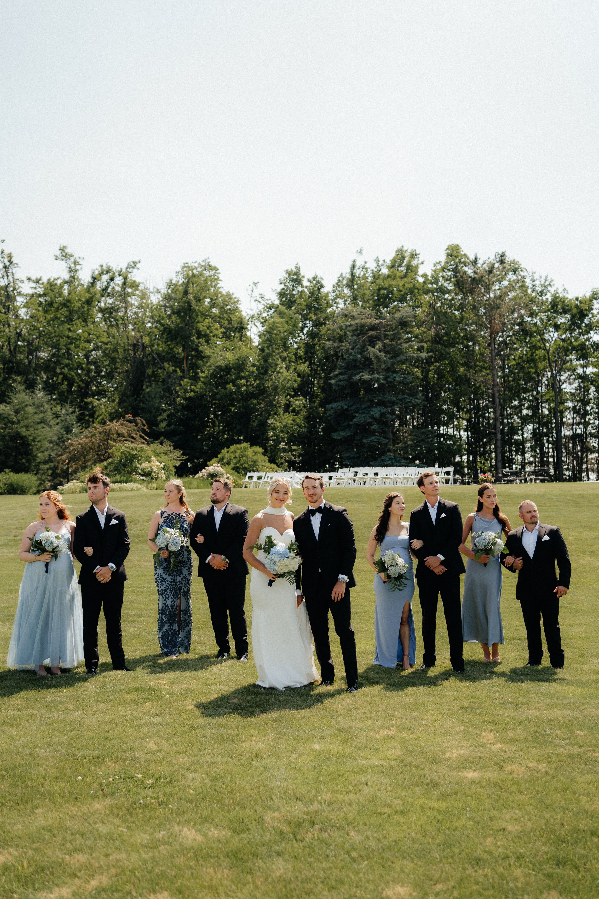 Wedding party standing in a line on green grass with bridesmaids in light blue dresses and groomsmen in black suits.