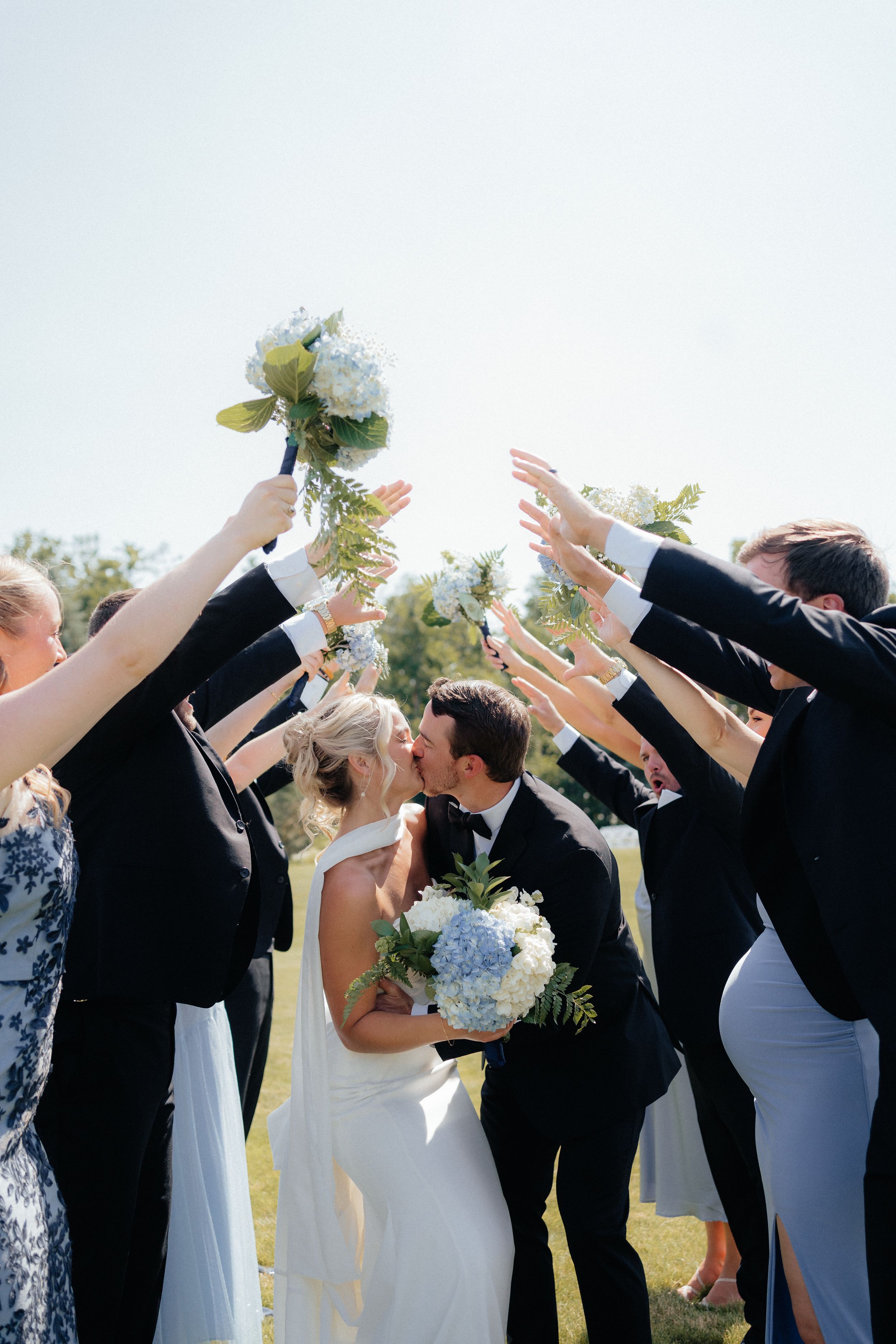 Wedding couple shares a kiss under an arch of raised arms and flowers during their joyful outdoor ceremony celebration.