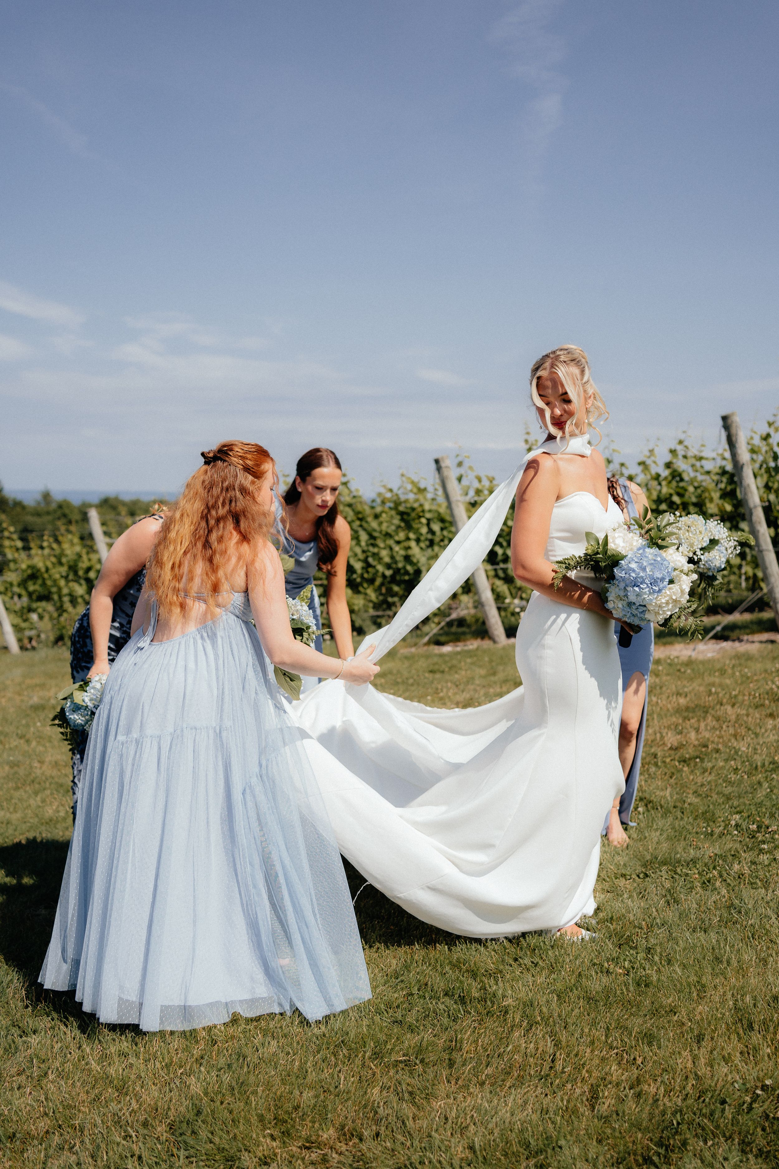 Bridesmaids in light blue dresses help arrange the flowing train of a white wedding gown on sunny grassy hillside.