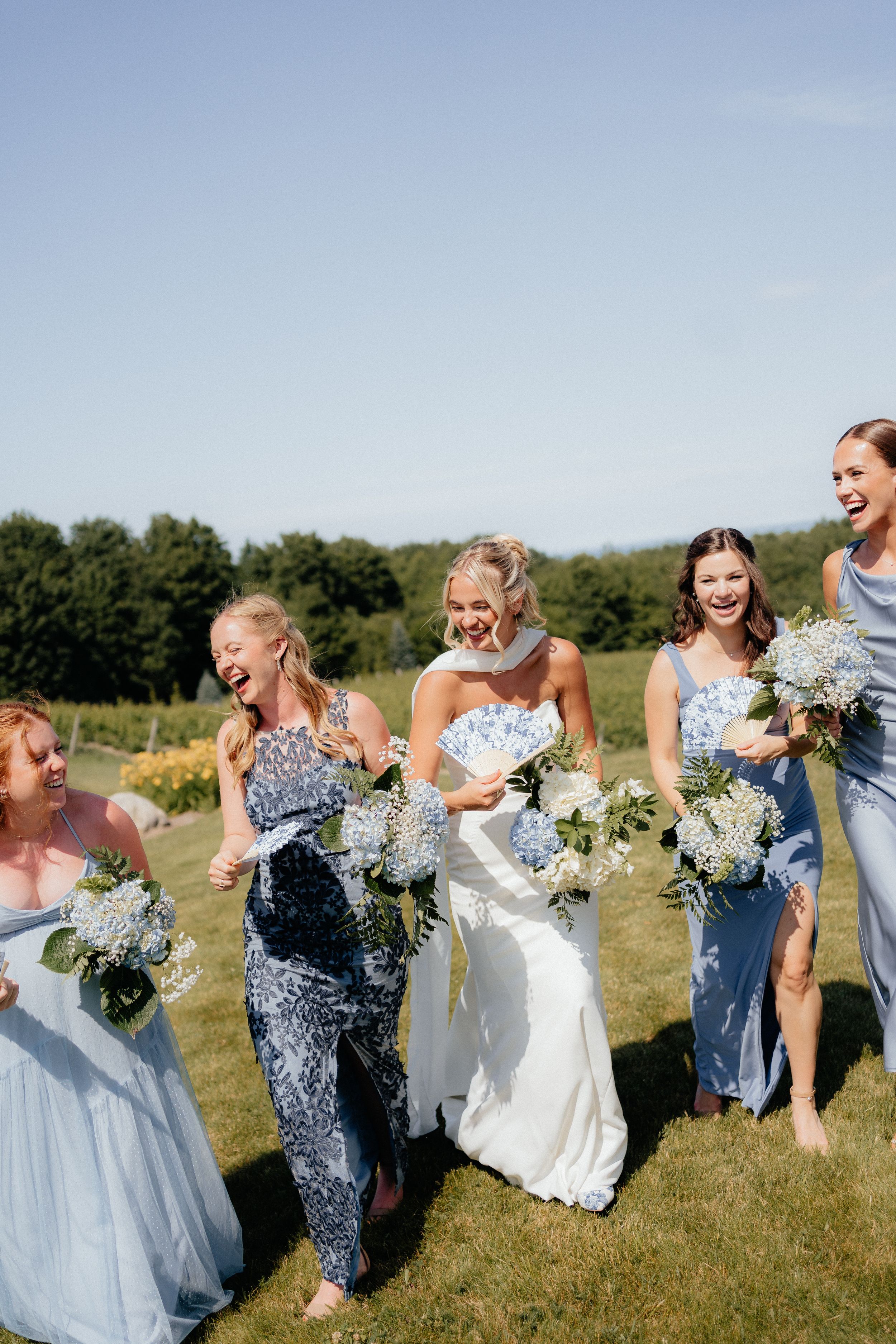 A bridal party laughs together while holding white and blue floral bouquets on a sunny outdoor lawn.