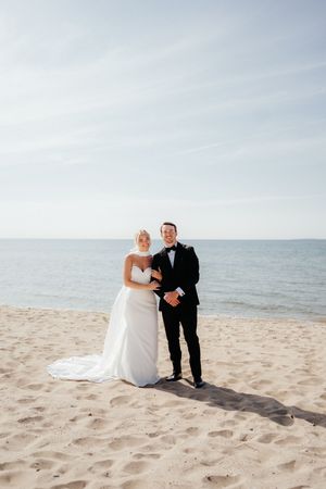 Newlywed couple poses together on a scenic beach with ocean waves and blue sky in the background.