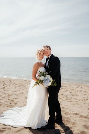 Elegant couple embraces on a sandy beach with blue hydrangea bouquet and ocean horizon behind them.