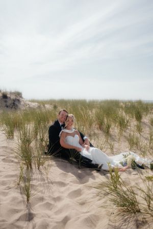 Couple in wedding attire lying romantically together on a sandy beach with tall grass in the background.