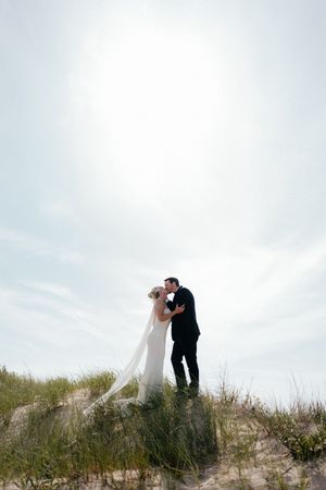 A romantic wedding couple embraces on a sandy dune overlooking a cloudy sky.