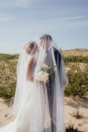 A flowing white veil drapes over a couple holding a bouquet of white flowers on sandy dunes.