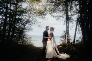 Wedding couple shares an intimate embrace among trees along a misty waterfront at dusk.