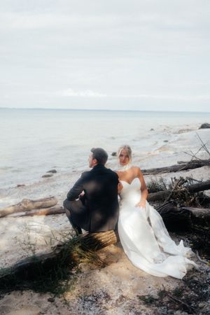 A couple in wedding attire sits on driftwood at a rocky beach shoreline during an overcast day.
