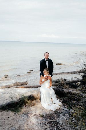 Couple in wedding attire poses on rocky beach shore with ocean in background during outdoor coastal photoshoot.
