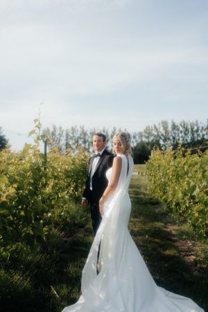 A couple in formal wedding attire poses together in a scenic vineyard at sunset.