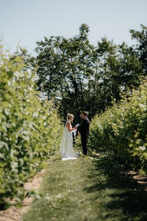 Two people in formal wear share an intimate moment on a garden path surrounded by tall bushes and trees.