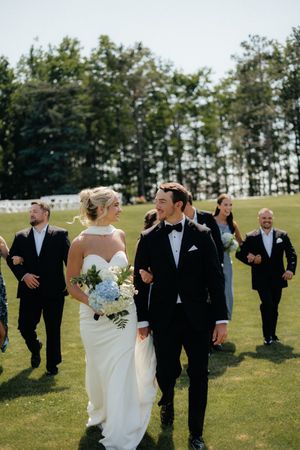 Wedding party walking and laughing together on a sunny day across a green lawn with trees in the background.
