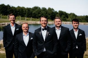 Groomsmen in black suits and white shirts posing together outdoors on a sunny day.