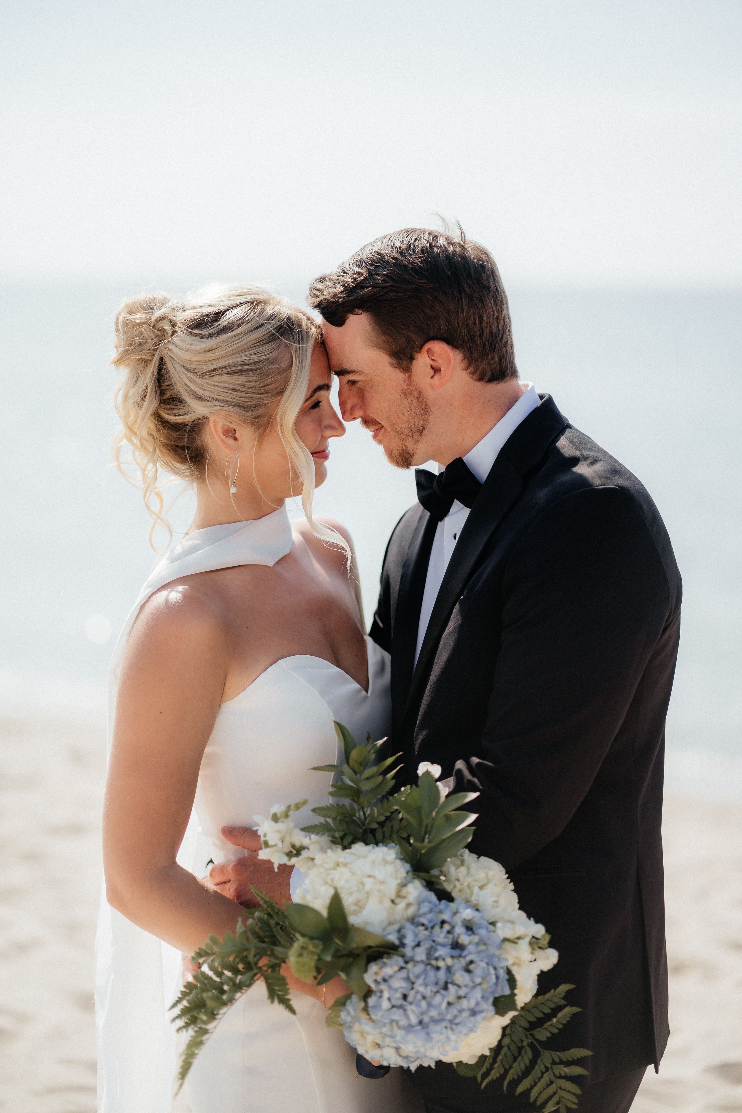 A romantic beachside wedding moment with the bride holding a blue and white hydrangea bouquet with greenery.