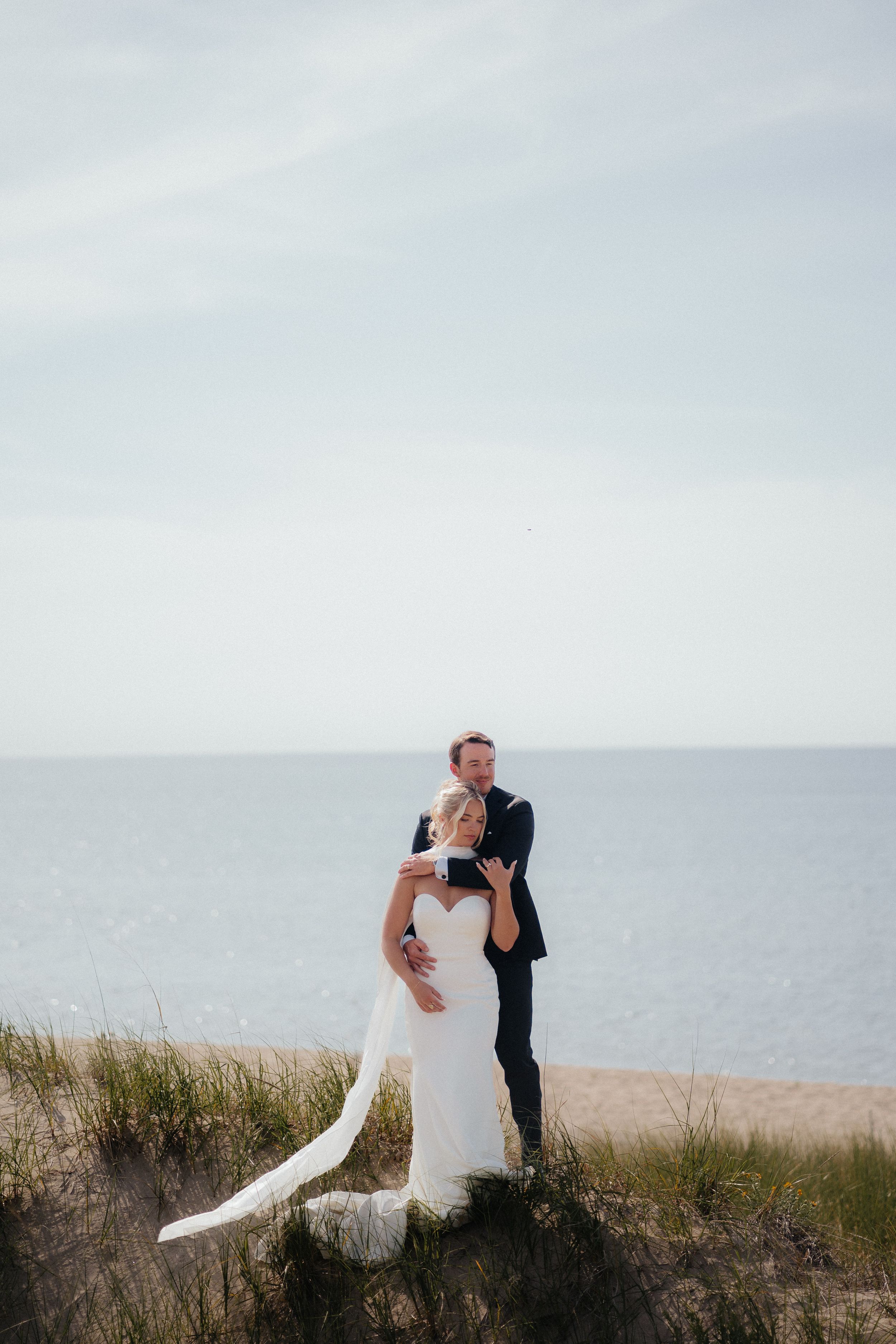 A couple embraces on a coastal clifftop overlooking the ocean with a cloudy sky in the background.