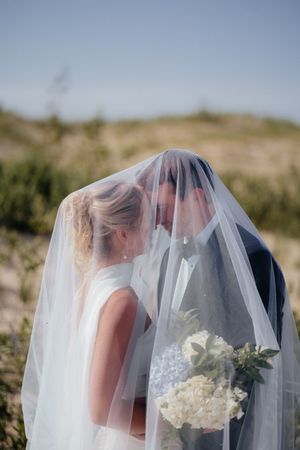 A romantic wedding moment captured under a flowing white veil with a bouquet of white flowers against a hillside backdrop.