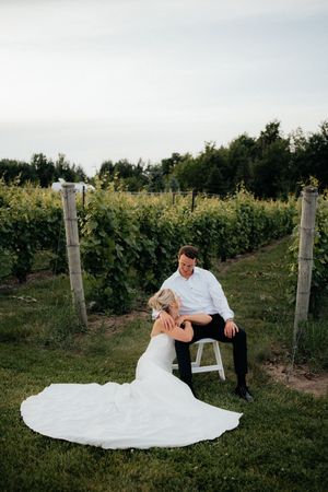 A couple shares an intimate moment in a vineyard with a flowing white wedding dress spread on the grass.