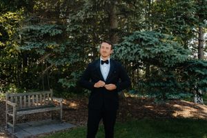 A well-dressed person in a black tuxedo and bowtie stands in front of evergreen trees.