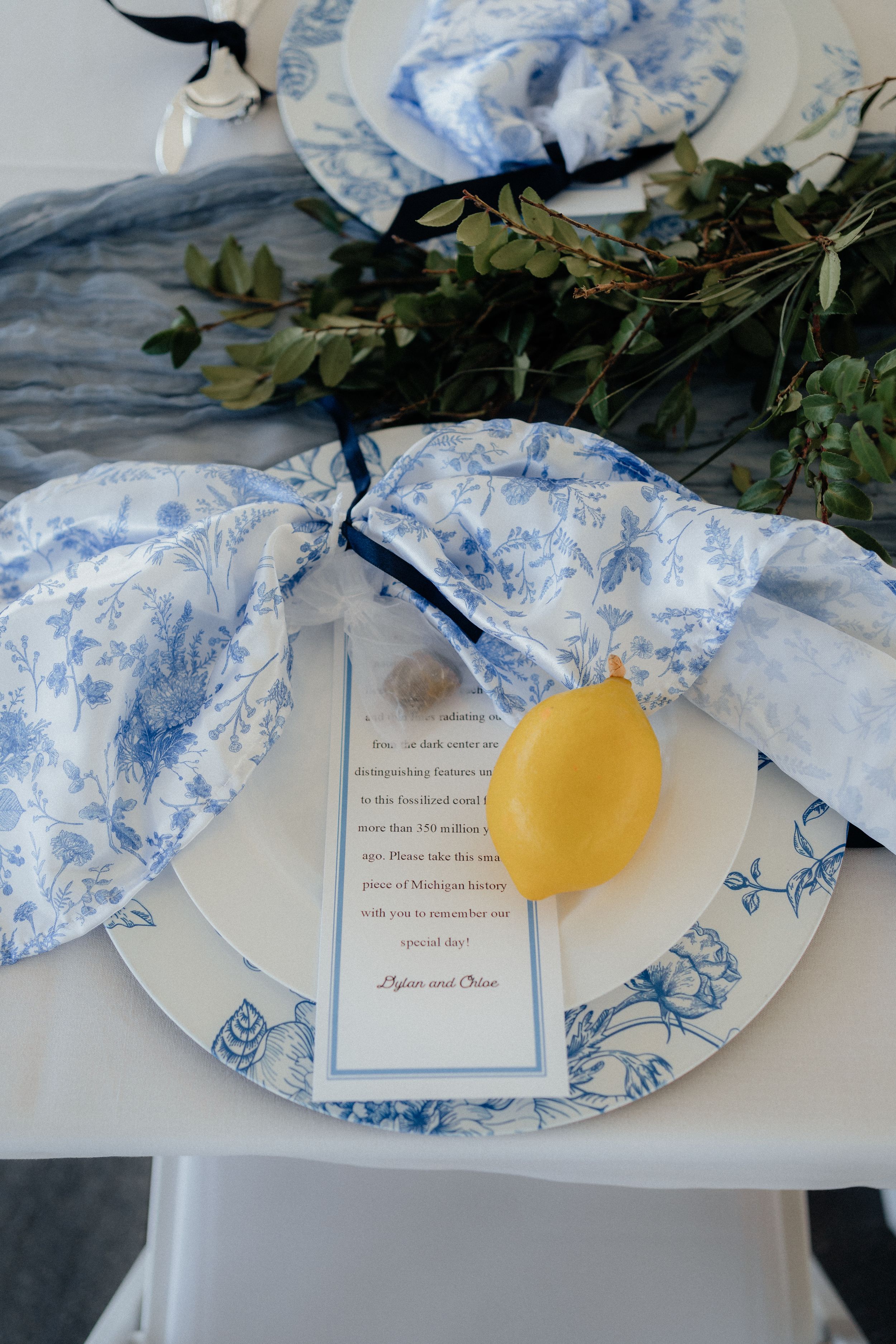 A place setting with blue and white floral china, a fresh lemon, and a menu card decorated with greenery.