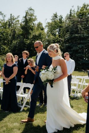 Wedding ceremony taking place outdoors on a sunny day with white chairs on green grass surrounded by trees.