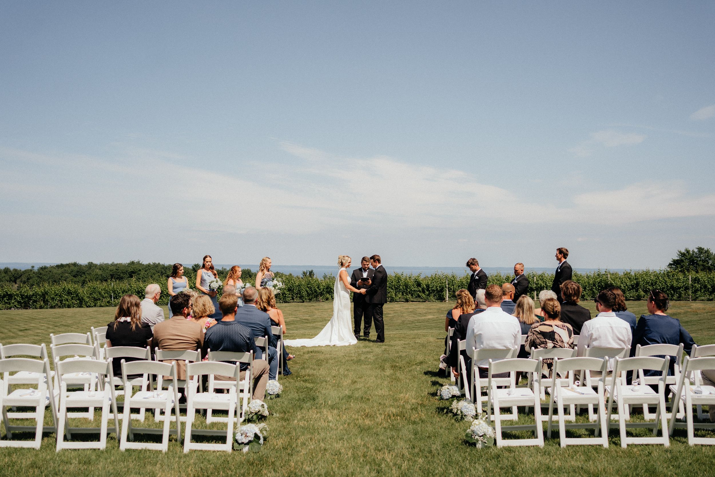 An outdoor wedding ceremony setup with white chairs on a grassy field overlooking scenic countryside views.