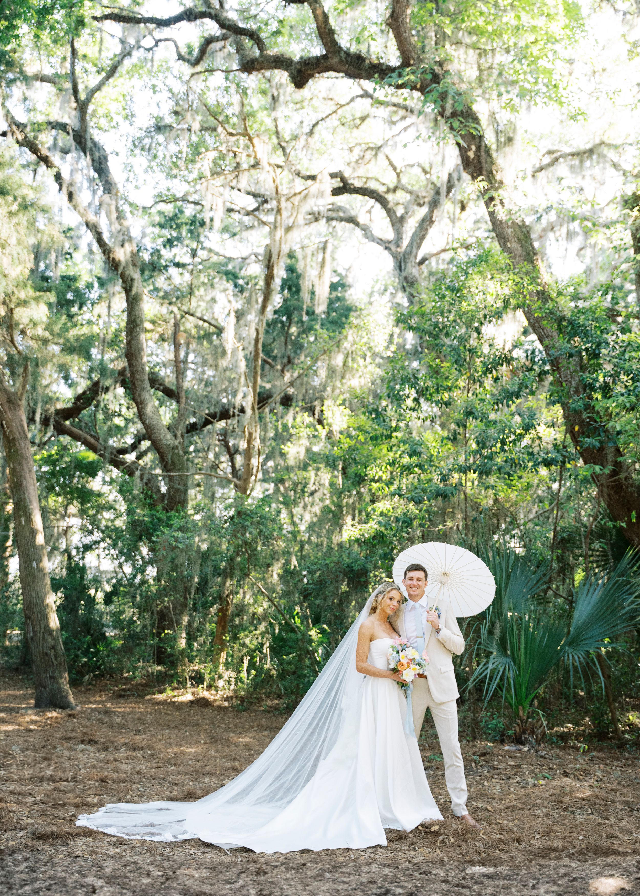 Bride and groom pose under Spanish moss-draped oak trees with a white parasol in natural light.