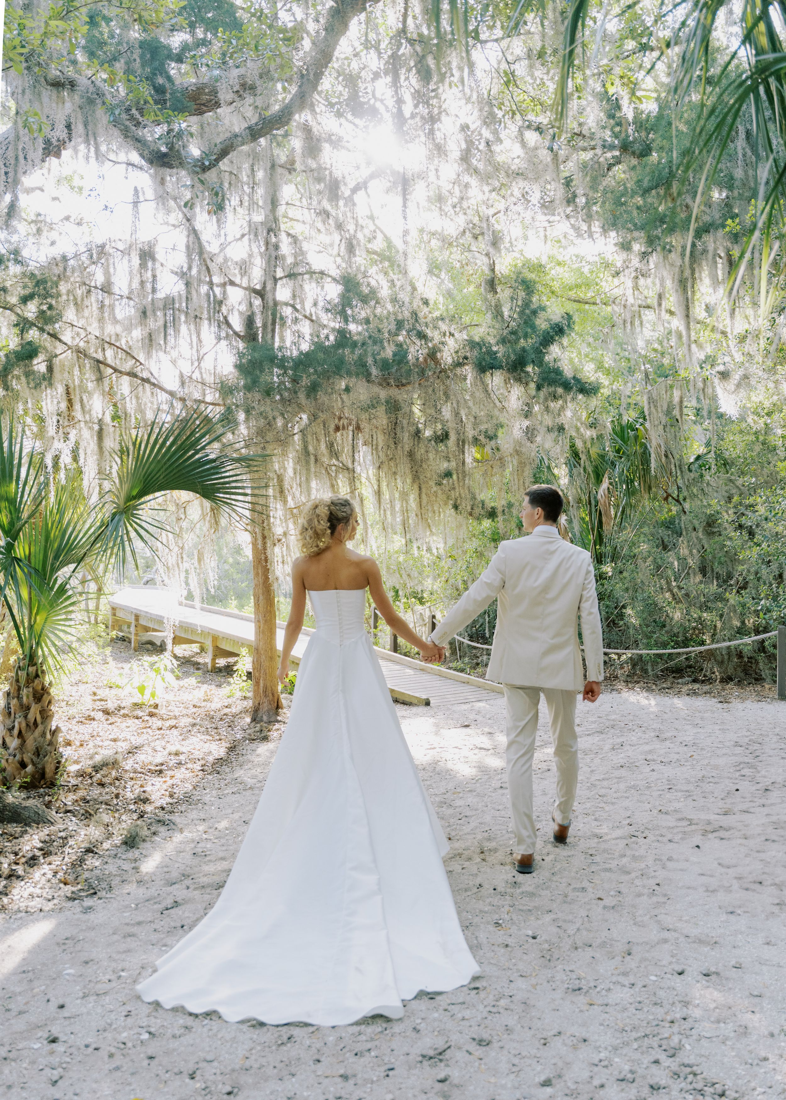 Bride and groom walk hand in hand through a sandy path beneath Spanish moss-draped trees.