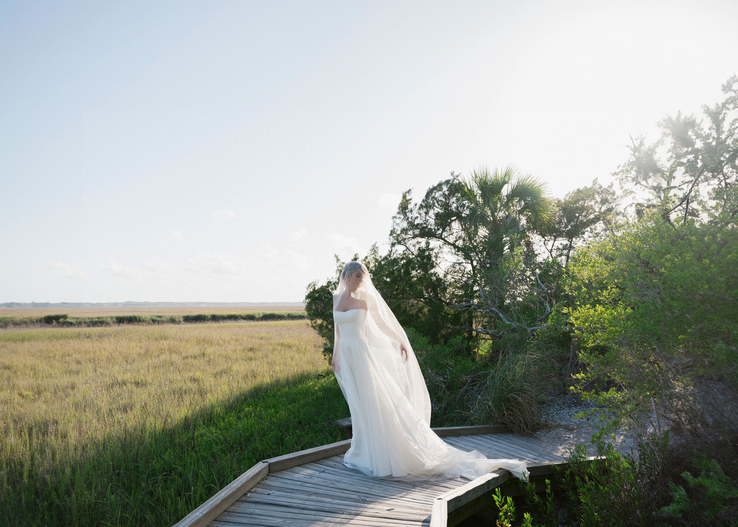 Bride in flowing white gown and veil stands on a wooden boardwalk overlooking a sunlit marsh.