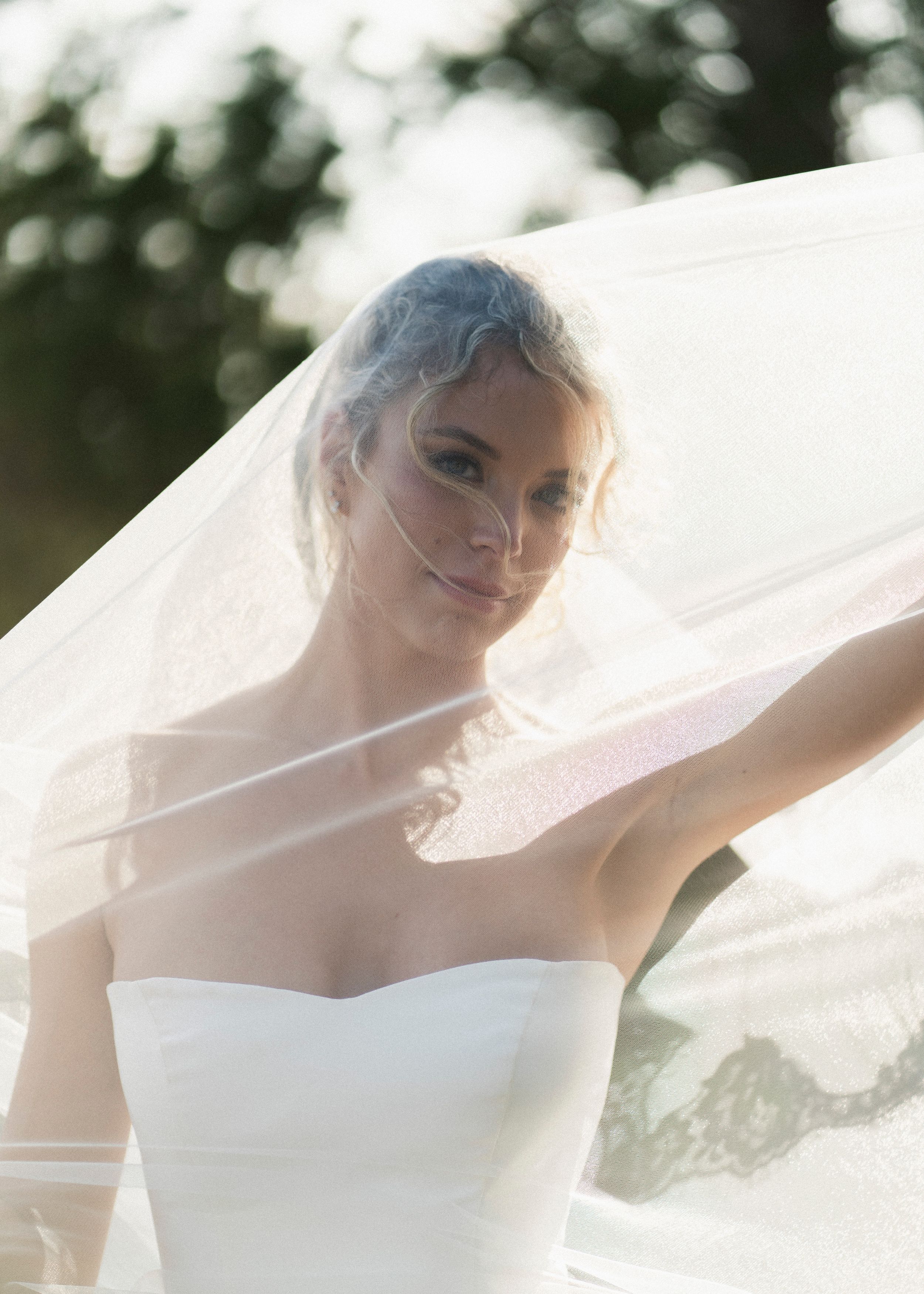 Radiant bride gazes through her flowing veil in soft natural light outdoors.