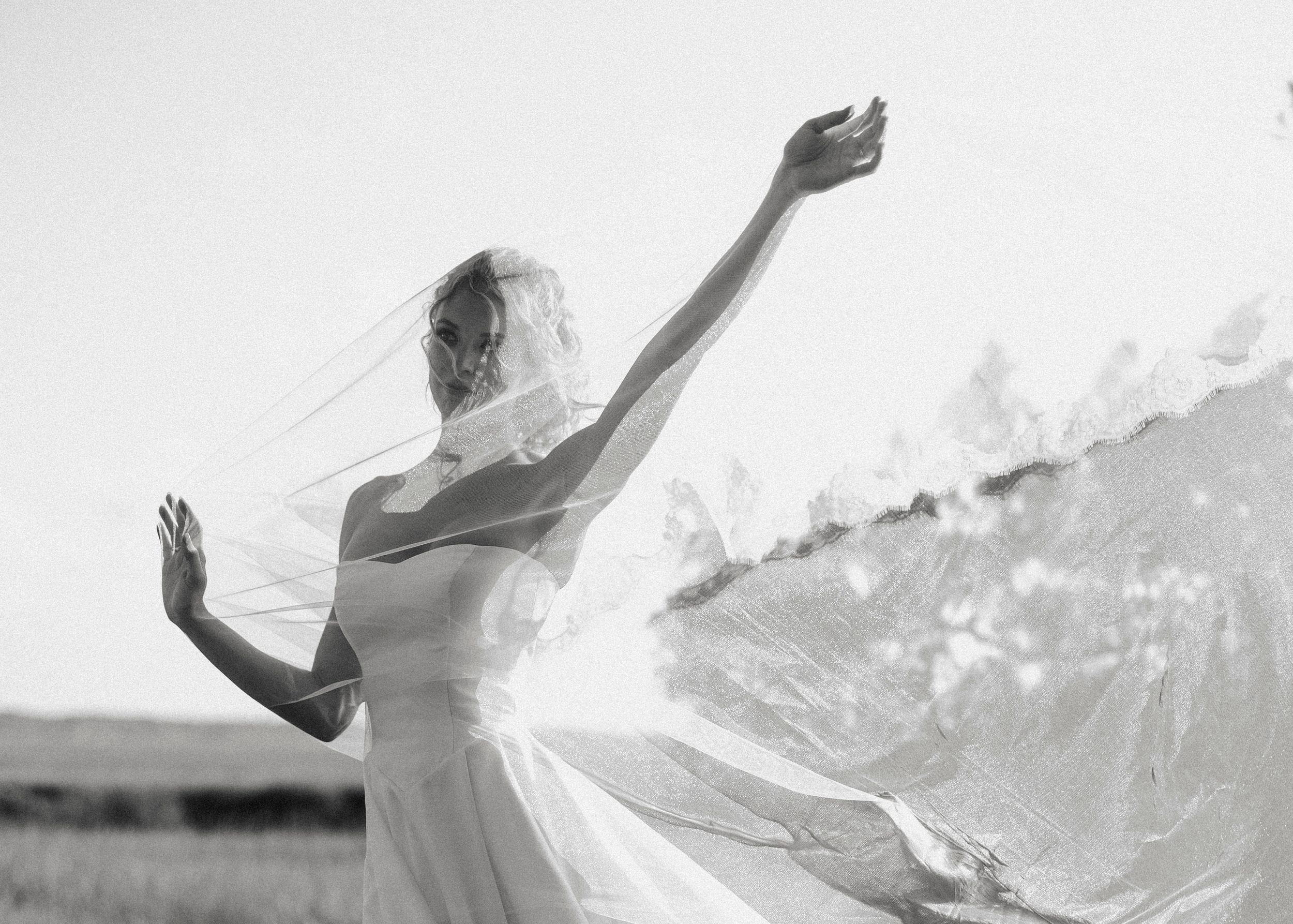 Black and white photo of a bride with veil flowing dramatically in the wind outdoors.