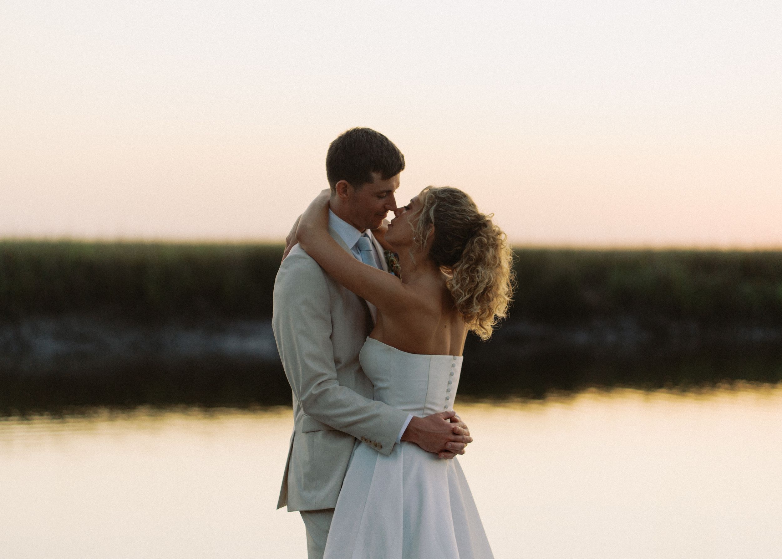 Bride and groom share a romantic embrace at golden hour beside a serene coastal waterway.
