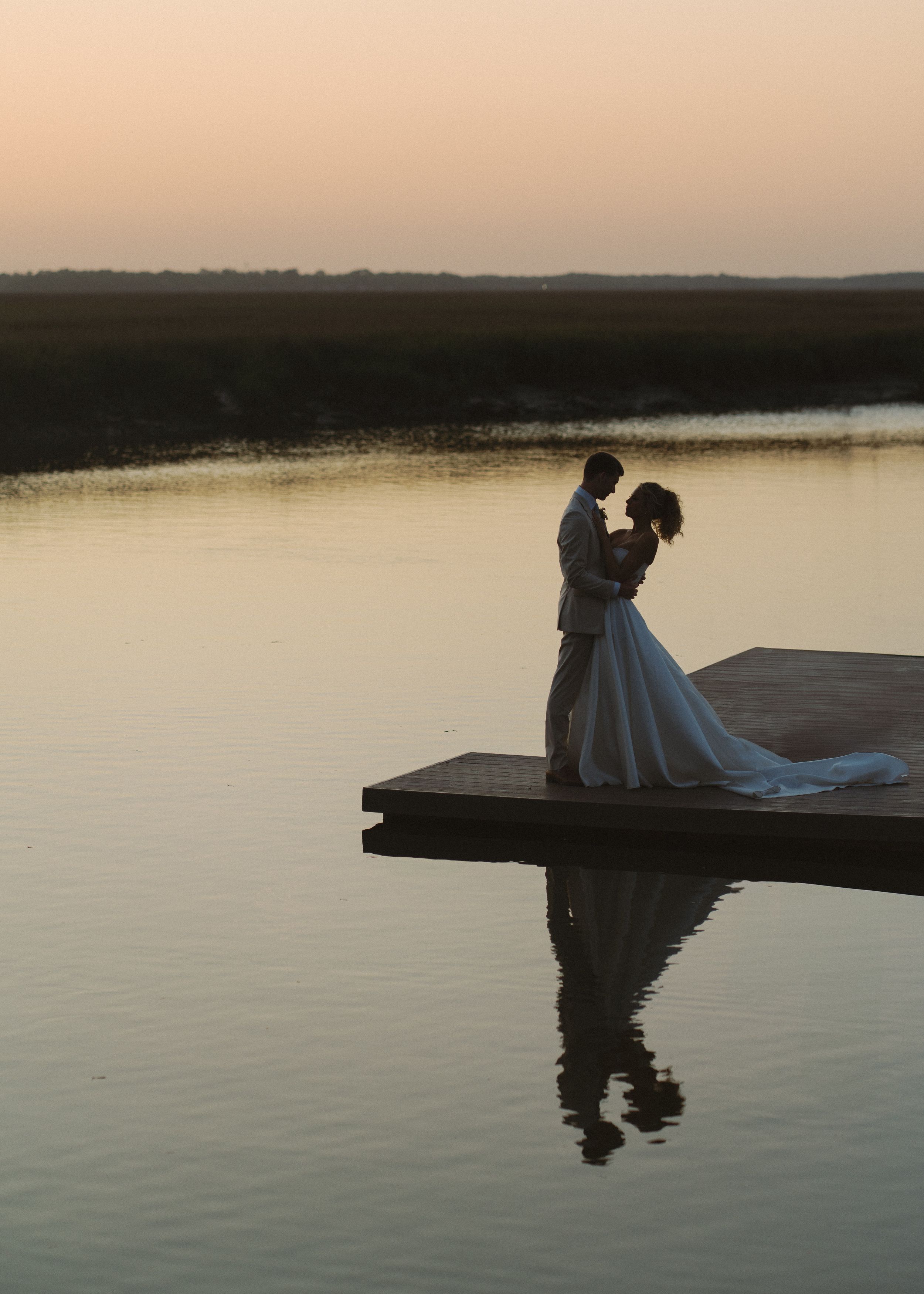 Bride and groom embrace on a dock reflected in calm water at dusk with a glowing horizon.