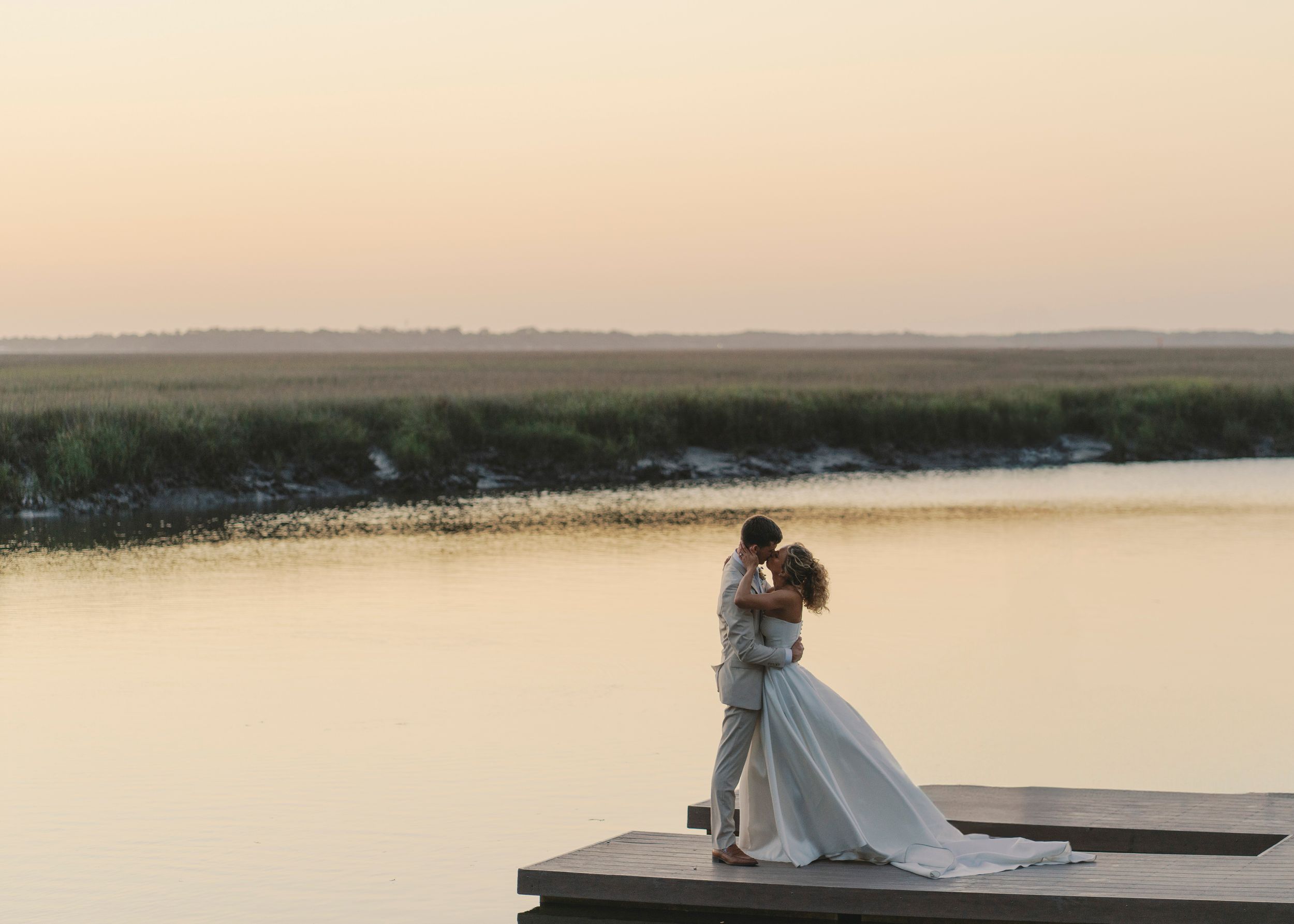 Bride and groom kiss on a wooden dock overlooking a tranquil marsh at sunset.