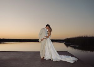 Bride and groom share a romantic embrace on a dock at sunset with calm water reflecting golden light.
