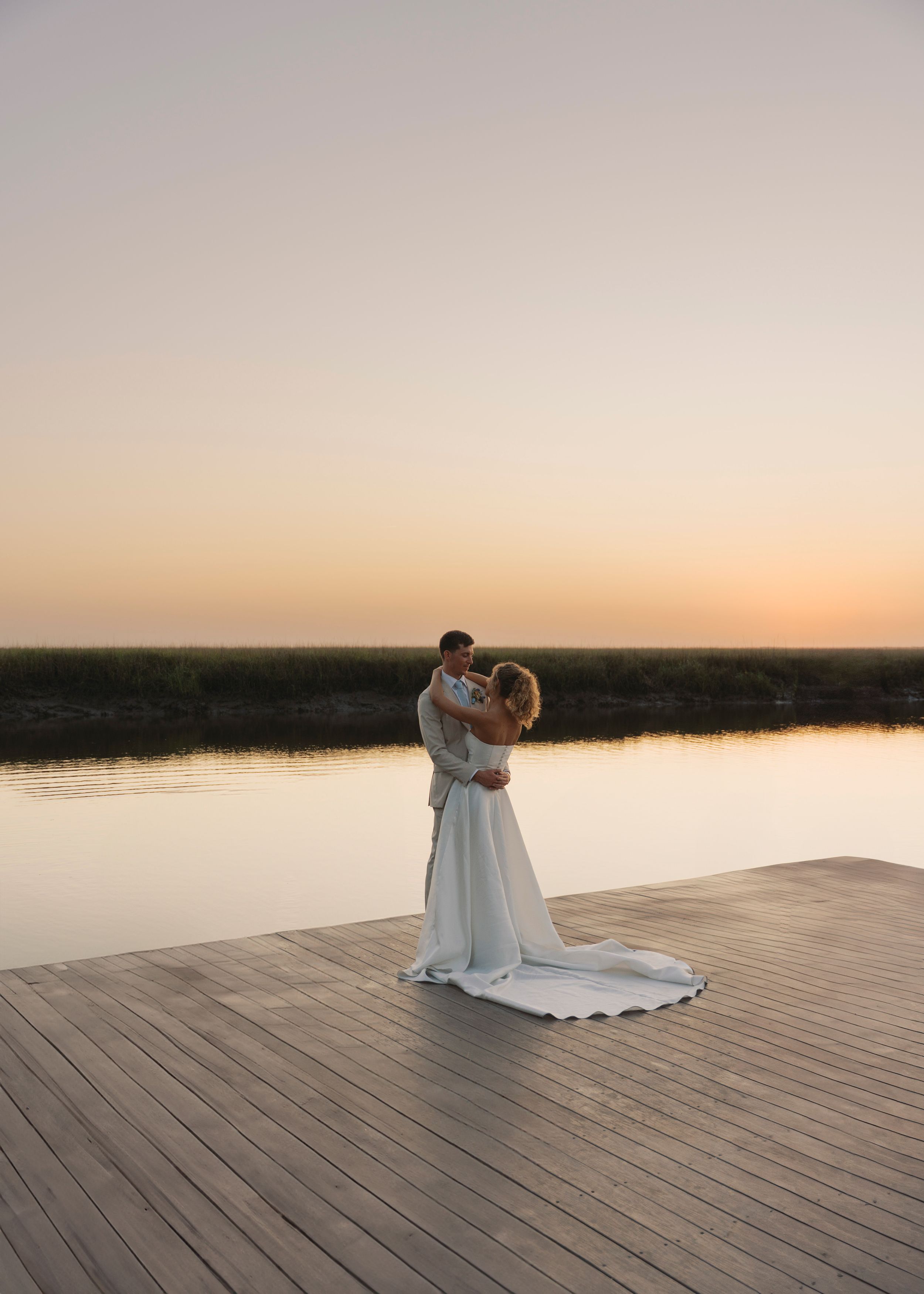 Wedding couple holds each other on a wooden dock during a serene golden hour sunset by the water.