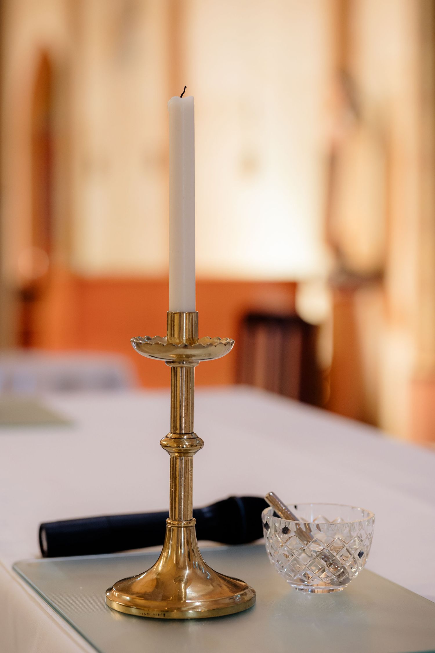 An elegant brass candlestick holder with a white taper candle sits next to a cut crystal bowl on a white tablecloth.