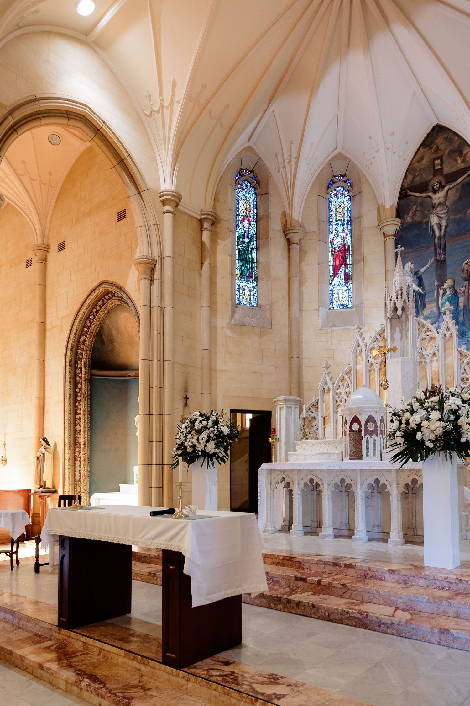 Interior of a Catholic church sanctuary with vaulted ceilings, stained glass windows, and white marble altar decorated with flowers.