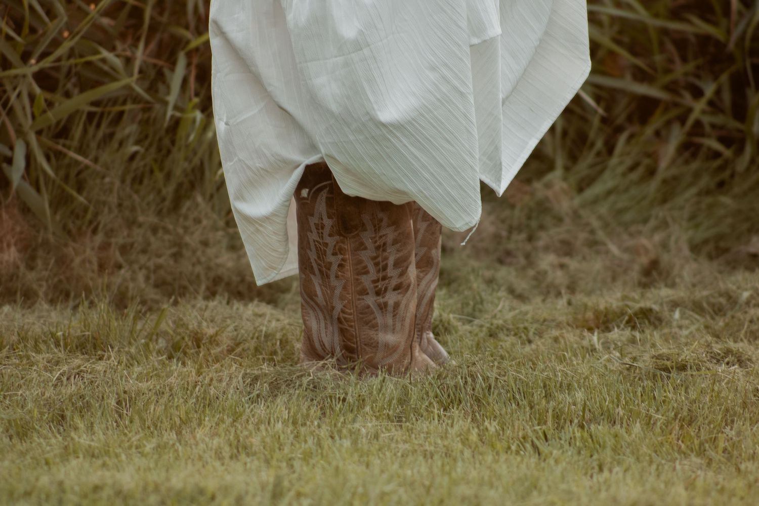 Brown leather boots peek out from under a white flowing dress against dewy grass.
