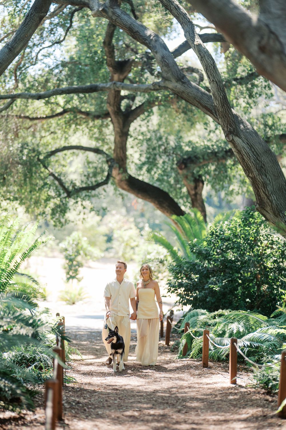 A couple in light colored outfits walking their dog along a scenic tree-lined path.