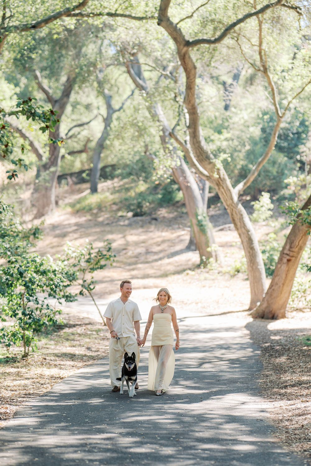 A couple walks their black dog down a sunlit wooded path in matching cream-colored outfits.