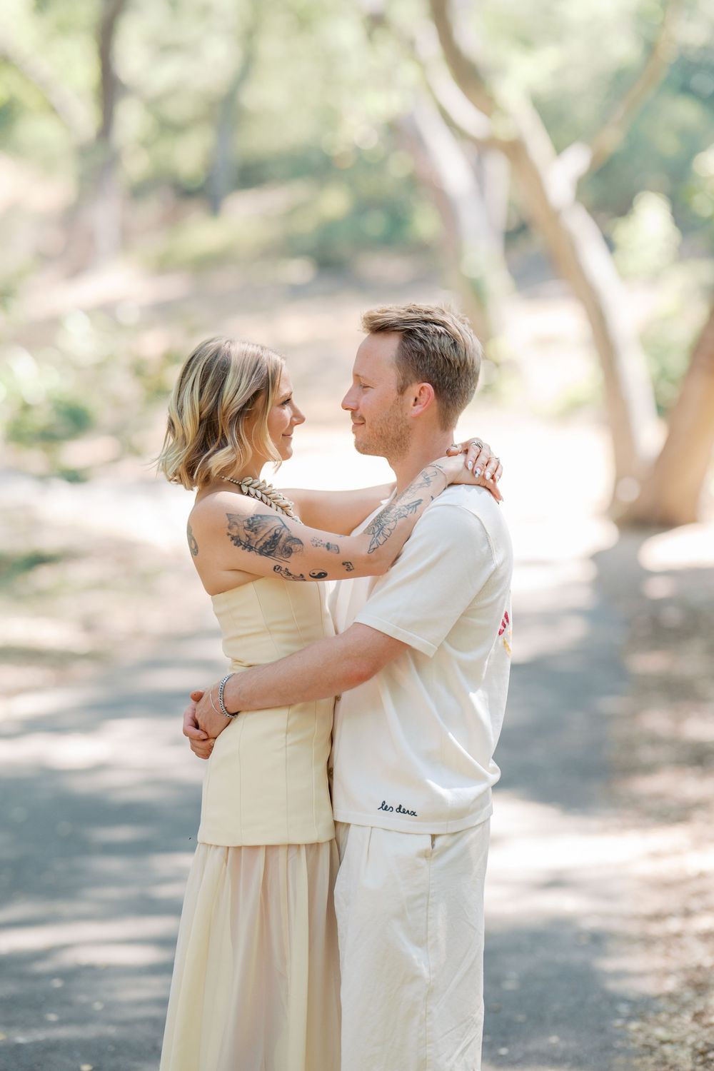 A romantic couple embraces on a sunlit nature trail wearing coordinating cream-colored outfits.