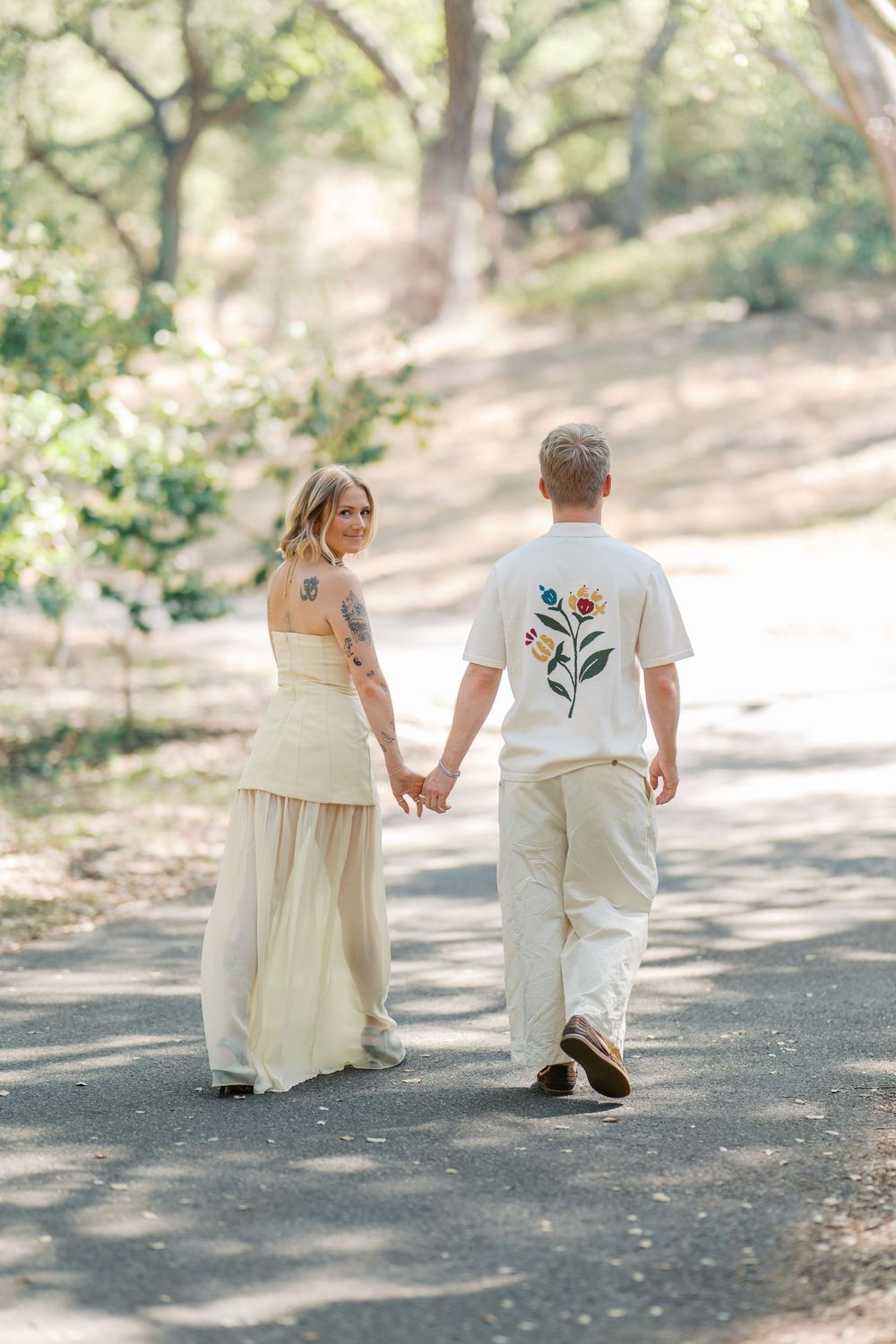 A couple holds hands while walking along a sunlit forest path in matching light-colored clothing.