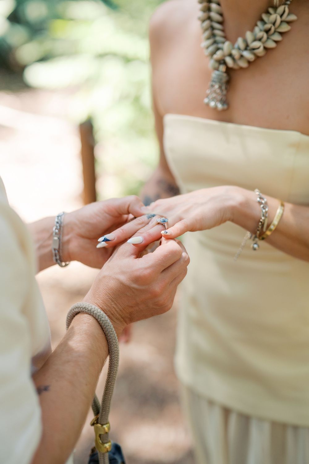 Close up of hands exchanging rings during an outdoor ceremony, with elegant jewelry and accessories visible.