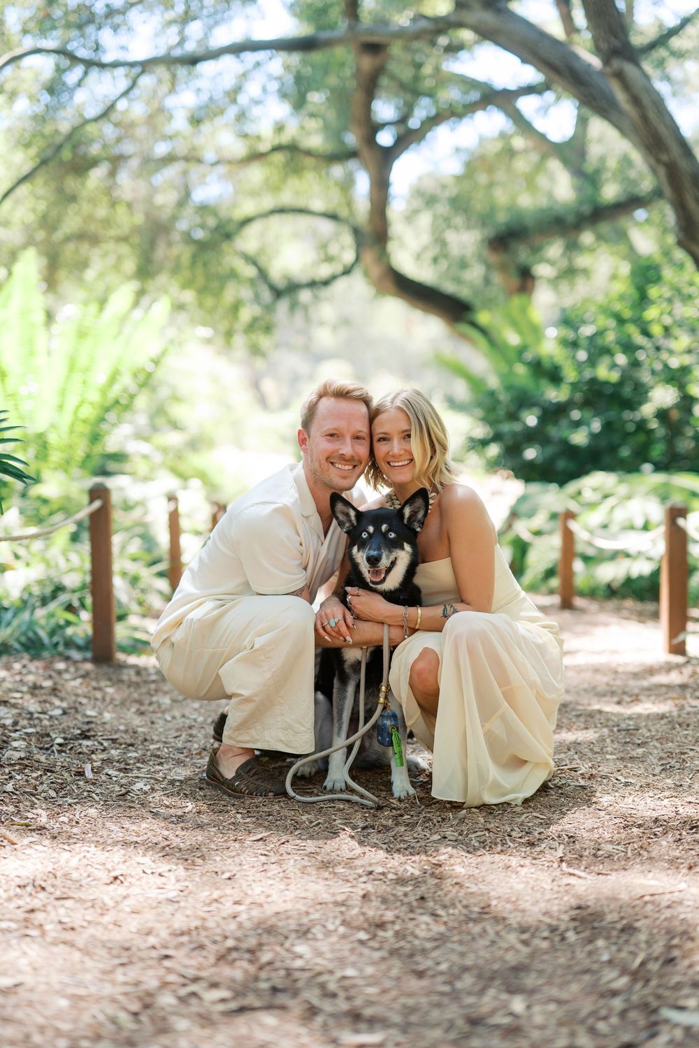 A couple relaxes on a forest floor with their black dog amidst dappled sunlight.