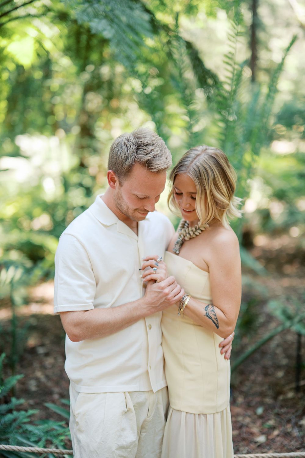 Romantic moment between couple in coordinating cream colored outfits surrounded by green forest foliage.
